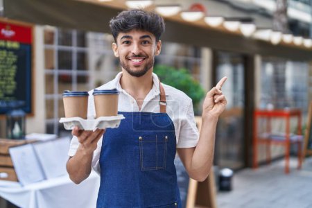 Téléchargez les photos : Arabe avec barbe portant tablier de serveur à la terrasse du restaurant souriant heureux pointant avec la main et le doigt sur le côté - en image libre de droit