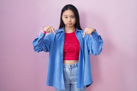 Foto de Young asian woman standing over pink background pointing down looking sad and upset, indicating direction with fingers, unhappy and depressed. - Imagen libre de derechos