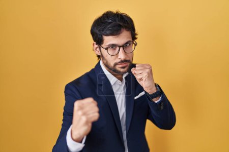 Foto de Handsome latin man standing over yellow background ready to fight with fist defense gesture, angry and upset face, afraid of problem - Imagen libre de derechos