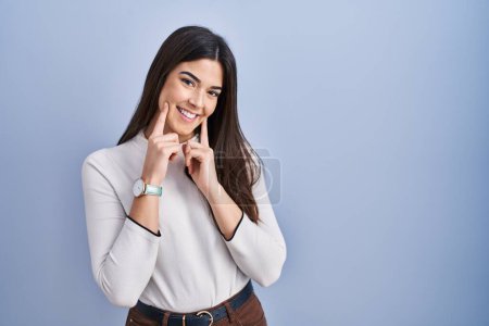 Photo for Young brunette woman standing over blue background smiling with open mouth, fingers pointing and forcing cheerful smile - Royalty Free Image