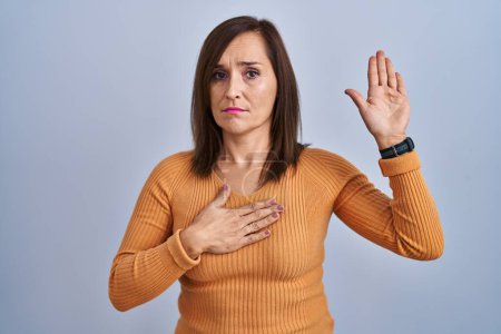 Photo for Middle age brunette woman standing wearing orange sweater swearing with hand on chest and open palm, making a loyalty promise oath - Royalty Free Image