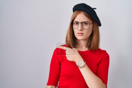 Photo for Young redhead woman standing wearing glasses and beret pointing aside worried and nervous with forefinger, concerned and surprised expression - Royalty Free Image