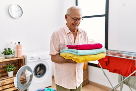 Téléchargez les photos : Senior homme souriant confiant tenant serviettes pliées à la buanderie - en image libre de droit