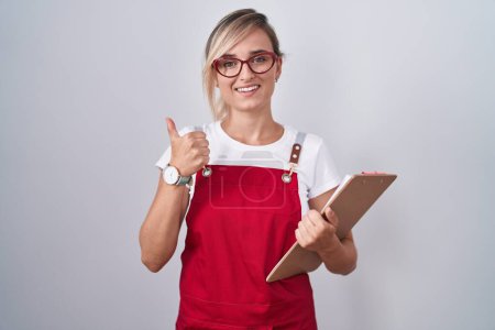 Photo for Young blonde woman wearing waiter uniform holding clipboard doing happy thumbs up gesture with hand. approving expression looking at the camera showing success. - Royalty Free Image