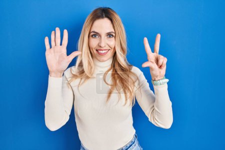 Foto de Joven mujer caucásica de pie sobre fondo azul mostrando y señalando hacia arriba con los dedos número ocho mientras sonríe confiado y feliz. - Imagen libre de derechos