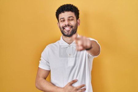 Foto de Hombre árabe de pie sobre fondo amarillo riéndose de ti, señalando con el dedo a la cámara con la mano sobre el cuerpo, expresión de vergüenza - Imagen libre de derechos