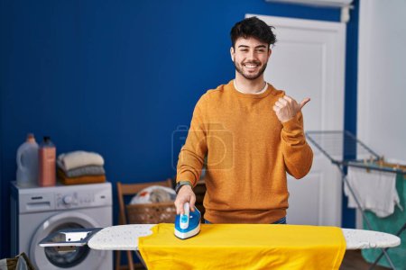 Foto de Hombre hispano con barba planchando ropa en casa apuntando el pulgar hacia el lado sonriendo feliz con la boca abierta - Imagen libre de derechos