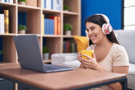 Téléchargez les photos : Young beautiful hispanic woman using laptop drinking coffee sitting on floor at home - en image libre de droit