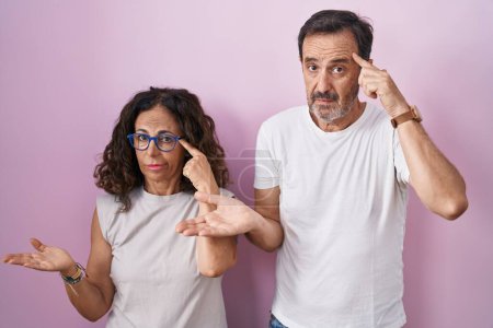 Photo for Middle age hispanic couple together over pink background confused and annoyed with open palm showing copy space and pointing finger to forehead. think about it. - Royalty Free Image