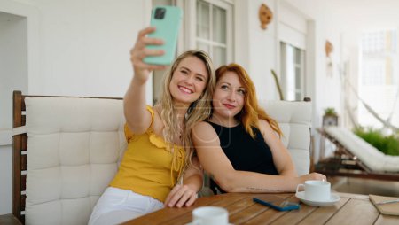 Photo for Two women make selfie by smartphone sitting on table at home terrace - Royalty Free Image