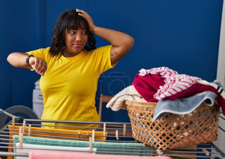Téléchargez les photos : Young beautiful latin woman looking clock hanging clothes on clothesline at laundry room - en image libre de droit