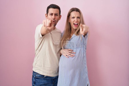 Téléchargez les photos : Jeune couple s'attendant à un bébé debout sur fond rose pointant mécontent et frustré à la caméra, en colère et furieux contre vous - en image libre de droit