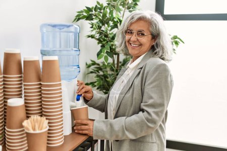 Téléchargez les photos : Femme d'affaires aux cheveux gris d'âge moyen souriant eau potable heureuse au bureau. - en image libre de droit