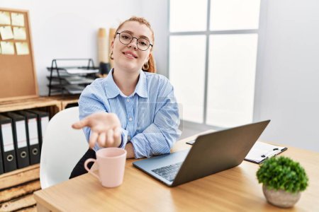 Photo for Young redhead woman working at the office using computer laptop smiling cheerful offering palm hand giving assistance and acceptance. - Royalty Free Image