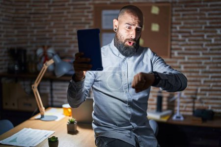 Téléchargez les photos : Jeune homme hispanique avec barbe et tatouages travaillant au bureau la nuit en regardant l'heure de la montre inquiet, peur de se retrouver en retard - en image libre de droit