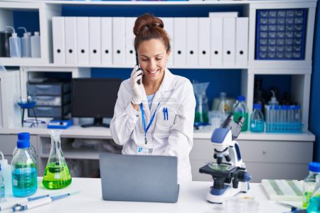Téléchargez les photos : Jeune femme scientifique parlant sur le smartphone en utilisant un ordinateur portable au laboratoire - en image libre de droit