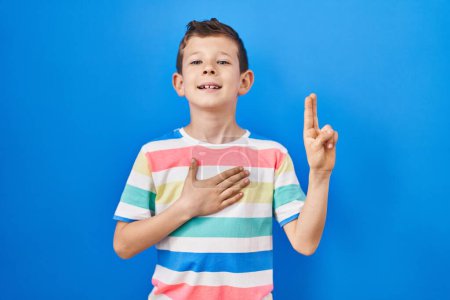 Photo for Young caucasian kid standing over blue background smiling swearing with hand on chest and fingers up, making a loyalty promise oath - Royalty Free Image