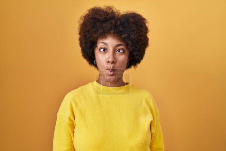 Téléchargez les photos : Jeune femme afro-américaine debout sur fond jaune faisant visage de poisson avec des lèvres, geste fou et comique. expression drôle. - en image libre de droit