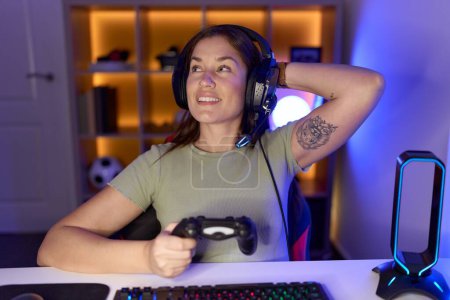 Foto de Hermosa mujer morena jugando videojuegos con auriculares sonriendo confiado tocar el pelo con la mano hacia arriba gesto, posando atractivo y de moda - Imagen libre de derechos