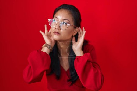 Photo for Asian young woman standing over red background trying to hear both hands on ear gesture, curious for gossip. hearing problem, deaf - Royalty Free Image