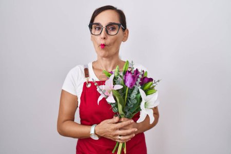 Photo for Middle age brunette woman wearing apron working at florist shop holding bouquet making fish face with lips, crazy and comical gesture. funny expression. - Royalty Free Image