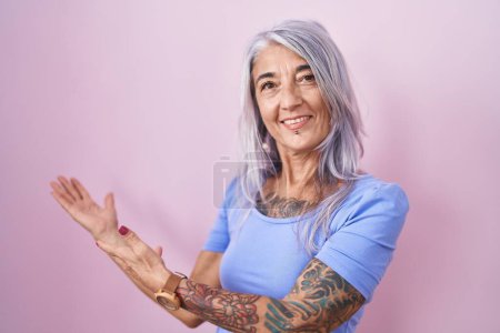 Téléchargez les photos : Femme d'âge moyen avec des tatouages debout sur fond rose invitant à entrer souriant naturel avec la main ouverte - en image libre de droit