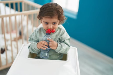 Foto de Adorable chica hispana sentada en una silla de bebé bebiendo agua en el dormitorio - Imagen libre de derechos