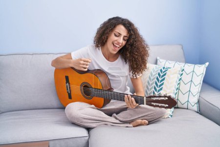 Téléchargez les photos : Jeune belle femme hispanique jouant de la guitare classique assise sur un canapé à la maison - en image libre de droit