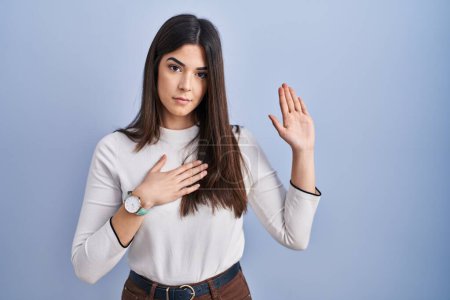 Photo for Young brunette woman standing over blue background swearing with hand on chest and open palm, making a loyalty promise oath - Royalty Free Image