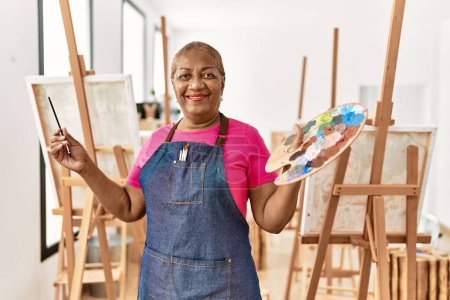 Téléchargez les photos : Femme afro-américaine senior souriant confiance tenant palette et pinceau au studio d'art - en image libre de droit
