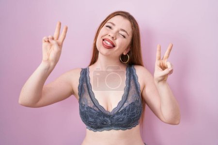 Photo for Redhead woman wearing lingerie over pink background smiling with tongue out showing fingers of both hands doing victory sign. number two. - Royalty Free Image