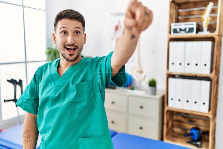 Photo for Young physiotherapist man working at pain recovery clinic pointing with finger surprised ahead, open mouth amazed expression, something on the front - Royalty Free Image