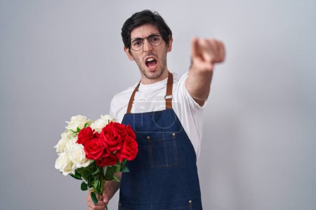 Foto de Young hispanic man holding bouquet of white and red roses pointing displeased and frustrated to the camera, angry and furious with you - Imagen libre de derechos