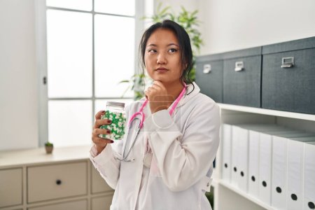 Foto de Joven asiática médico mujer celebración píldoras seria cara pensando en la pregunta con la mano en la barbilla, reflexivo acerca de la idea confusa - Imagen libre de derechos