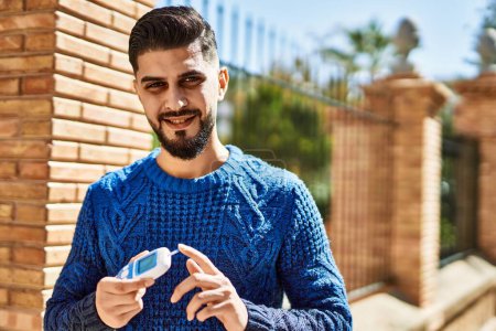 Téléchargez les photos : Jeune homme arabe souriant confiant tenant glucomètre à la rue - en image libre de droit