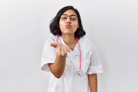 Photo for Young hispanic doctor woman wearing stethoscope over isolated background looking at the camera blowing a kiss with hand on air being lovely and sexy. love expression. - Royalty Free Image