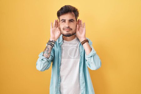 Photo for Young hispanic man with tattoos standing over yellow background trying to hear both hands on ear gesture, curious for gossip. hearing problem, deaf - Royalty Free Image