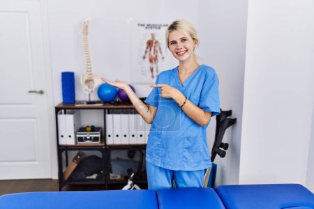 Foto de Mujer fisioterapeuta joven que trabaja en la clínica de recuperación del dolor sorprendida y sonriendo a la cámara mientras se presenta con la mano y señalando con el dedo. - Imagen libre de derechos