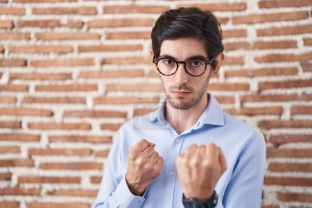 Foto de Young hispanic man standing over brick wall background ready to fight with fist defense gesture, angry and upset face, afraid of problem - Imagen libre de derechos