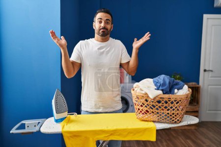 Téléchargez les photos : Young hispanic man with beard ironing clothes at home shouting and screaming loud to side with hand on mouth. communication concept. - en image libre de droit