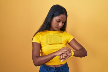 Téléchargez les photos : Young indian woman standing over yellow background checking the time on wrist watch, relaxed and confident - en image libre de droit