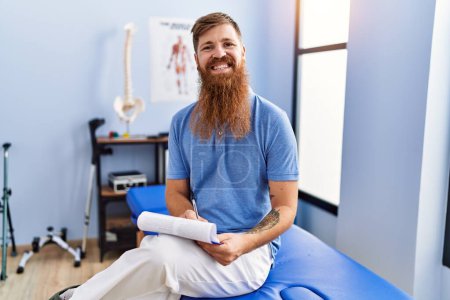 Photo for Young redhead man wearing physiotherapist uniform writing medical report at physiotherapy clinic - Royalty Free Image