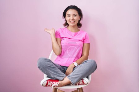 Photo for Hispanic young woman sitting on chair over pink background smiling cheerful presenting and pointing with palm of hand looking at the camera. - Royalty Free Image