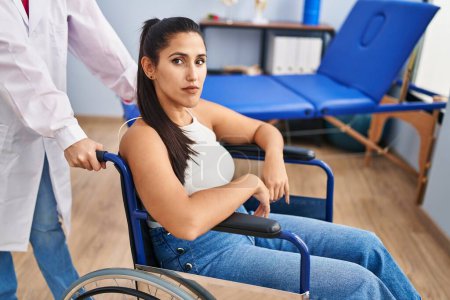 Photo for Young hispanic woman sitting on wheelchair at physiotherapy clinic thinking attitude and sober expression looking self confident - Royalty Free Image