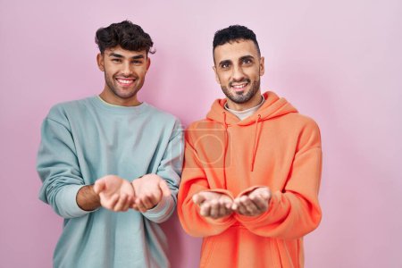 Foto de Joven pareja gay hispana de pie sobre fondo rosa sonriendo con las palmas de las manos juntas recibiendo o dando gesto. retención y protección - Imagen libre de derechos
