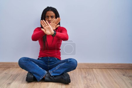 Foto de Young african american with braids sitting on the floor at home rejection expression crossing arms and palms doing negative sign, angry face - Imagen libre de derechos