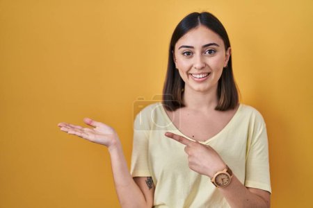Téléchargez les photos : Hispanic girl wearing casual t shirt over yellow background amazed and smiling to the camera while presenting with hand and pointing with finger. - en image libre de droit