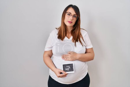 Photo for Pregnant woman holding baby ecography relaxed with serious expression on face. simple and natural looking at the camera. - Royalty Free Image