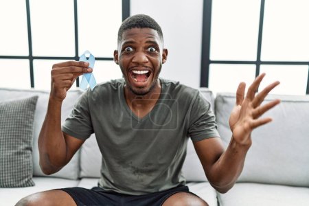 Foto de Young african american man holding blue ribbon sitting on the sofa at home celebrating victory with happy smile and winner expression with raised hands - Imagen libre de derechos
