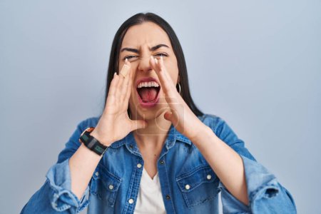 Photo for Hispanic woman standing over blue background shouting angry out loud with hands over mouth - Royalty Free Image